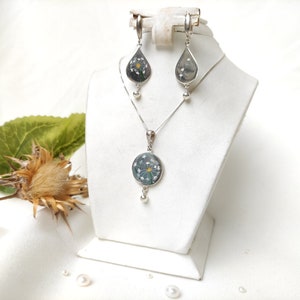May include: A silver necklace and earrings set with a delicate flower design encased in clear resin. The pendant is round and the earrings are teardrop shaped.