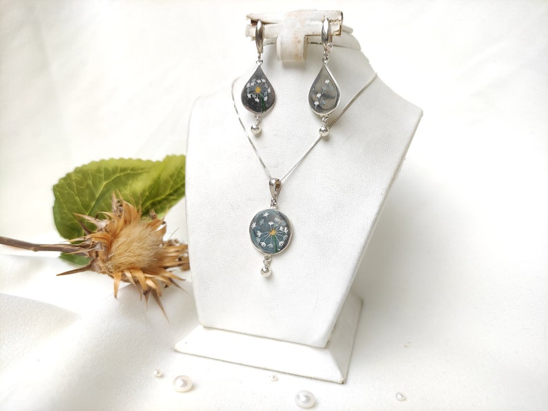 May include: A silver necklace and earrings set with a delicate flower design encased in clear resin. The pendant is round and the earrings are teardrop shaped.