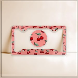 May include: A license plate frame and matching round emblem featuring a repeating pattern of red cherries with green leaves on a pink background. The frame has a rectangular shape with rounded corners and a central opening.