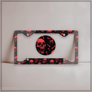 Puede incluir: Marco de matrícula negro con un patrón de calaveras rojas. El marco tiene un patrón repetido de calaveras y huesos rojos sobre un fondo negro. Un emblema circular en el centro presenta un diseño de calavera y hueso rojo.