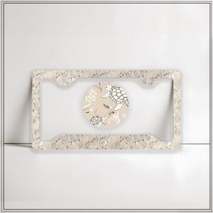 May include: Tan license plate frame with a repeating pattern of turtles and foliage. A circular insert features two turtles and plants. The design is in black and white on a tan background.