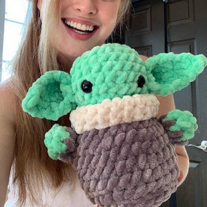 Peut inclure: Une femme tient un jouet en peluche crochet&eacute; vert et gris qui ressemble &agrave; un personnage de la franchise Star Wars. Le jouet a de grandes oreilles, de grands yeux et un col crochet&eacute; blanc.