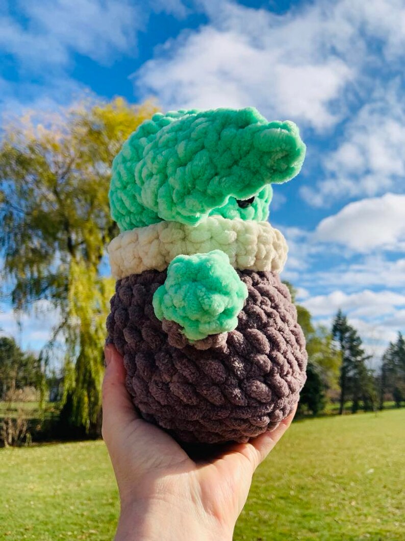 Peut inclure: Un jouet en peluche crochet&eacute; en forme de personnage extraterrestre vert. Le jouet a un corps brun, un col blanc et une t&ecirc;te verte avec de grands yeux. Le jouet est tenu dans la main d'une personne.