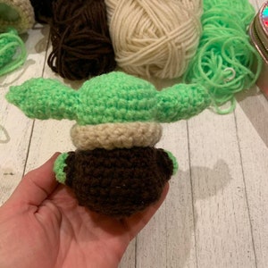 Może przedstawiać: Pluszowa zabawka Baby Yoda wykonana na szydełku w kolorze zielonym i brązowym z beżowym kapeluszem. Zabawka jest trzymana w dłoni.