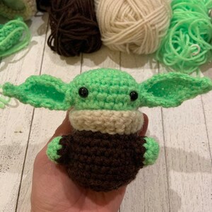 Może przedstawiać: Zielona pluszowa zabawka amigurumi wykonana na szydełku z brązowymi i beżowymi detalami. Zabawka ma duże oczy i przypomina popularną postać fikcyjną.