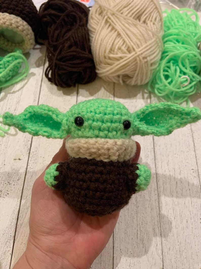 Może przedstawiać: Zielona pluszowa zabawka amigurumi wykonana na szydełku z brązowymi i beżowymi detalami. Zabawka ma duże oczy i przypomina popularną postać fikcyjną.
