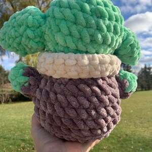 Peut inclure: Un jouet en peluche crochet&eacute; avec une t&ecirc;te verte, un col blanc et un corps brun. Le jouet est tenu dans une main.