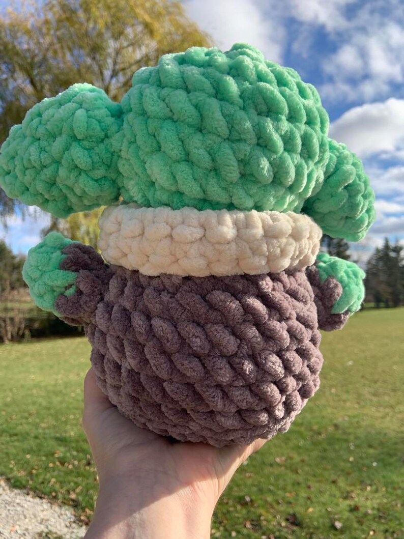 Peut inclure: Un jouet en peluche crochet&eacute; avec une t&ecirc;te verte, un col blanc et un corps brun. Le jouet est tenu dans une main.