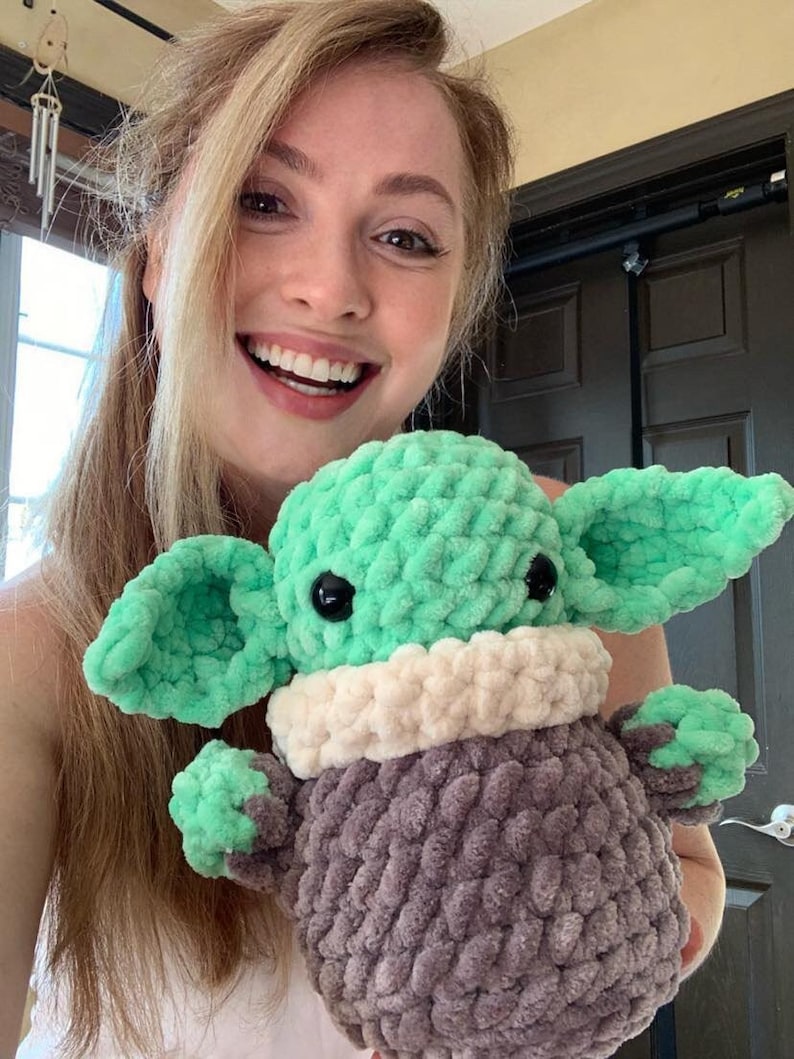 Peut inclure: Une femme tient un jouet en peluche crochet&eacute; vert et gris qui ressemble &agrave; un personnage de la franchise Star Wars. Le jouet a de grandes oreilles, de grands yeux et un col crochet&eacute; blanc.
