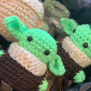 Może przedstawiać: Trzy pluszowe figurki Yody amigurumi wykonane na szydełku w kolorze zielonym i brązowym w czarnej torbie. Pluszaki mają białe akcenty i czarne oczy. Na torbie widnieje napis "Niech Moc będzie z Tobą!" wraz z zieloną emotką serca.