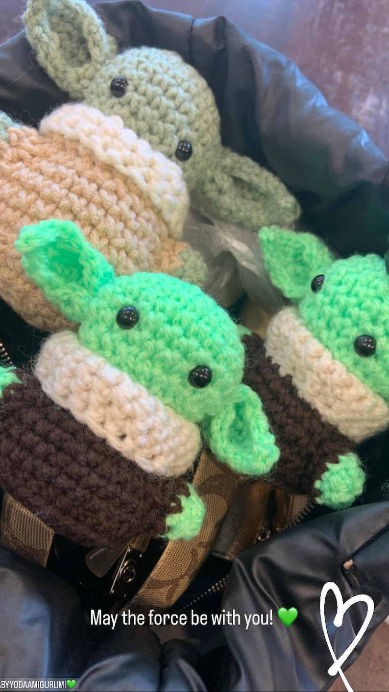 Może przedstawiać: Trzy pluszowe figurki Yody amigurumi wykonane na szydełku w kolorze zielonym i brązowym w czarnej torbie. Pluszaki mają białe akcenty i czarne oczy. Na torbie widnieje napis "Niech Moc będzie z Tobą!" wraz z zieloną emotką serca.