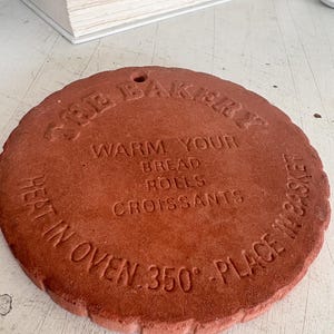 May include: A round, terracotta-colored ceramic bread warmer with the text "THE BAKERY" and instructions to warm bread, rolls, and croissants at 350 degrees Fahrenheit. The edge is scalloped.