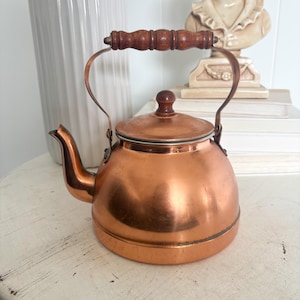 May include: A polished copper tea kettle with a rounded body, a curved spout, and a matching copper lid featuring a small wooden knob. The kettle includes a hinged metal handle topped with a dark, turned wooden grip for easy handling.