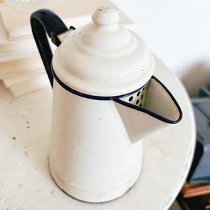 May include: White enamelware coffee pot with a dark blue handle and trim. The pot has a lid with a small knob and a spout. The coffee pot is sitting on a white surface.