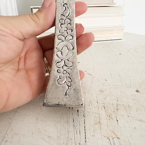 May include: A vintage silver metal tool with a floral design. The tool is rectangular with a tapered shape and features embossed flowers and leaves. The metal has a worn, antique appearance. The tool is held in a hand.