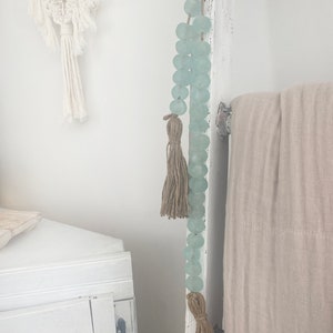 Op de afbeelding: Een lichtblauwe glazen kralenkrans met bruine kwastjes die aan een witte houten ladder hangt. De krans is gemaakt van ronde kralen en heeft een rustieke, bohemian stijl.