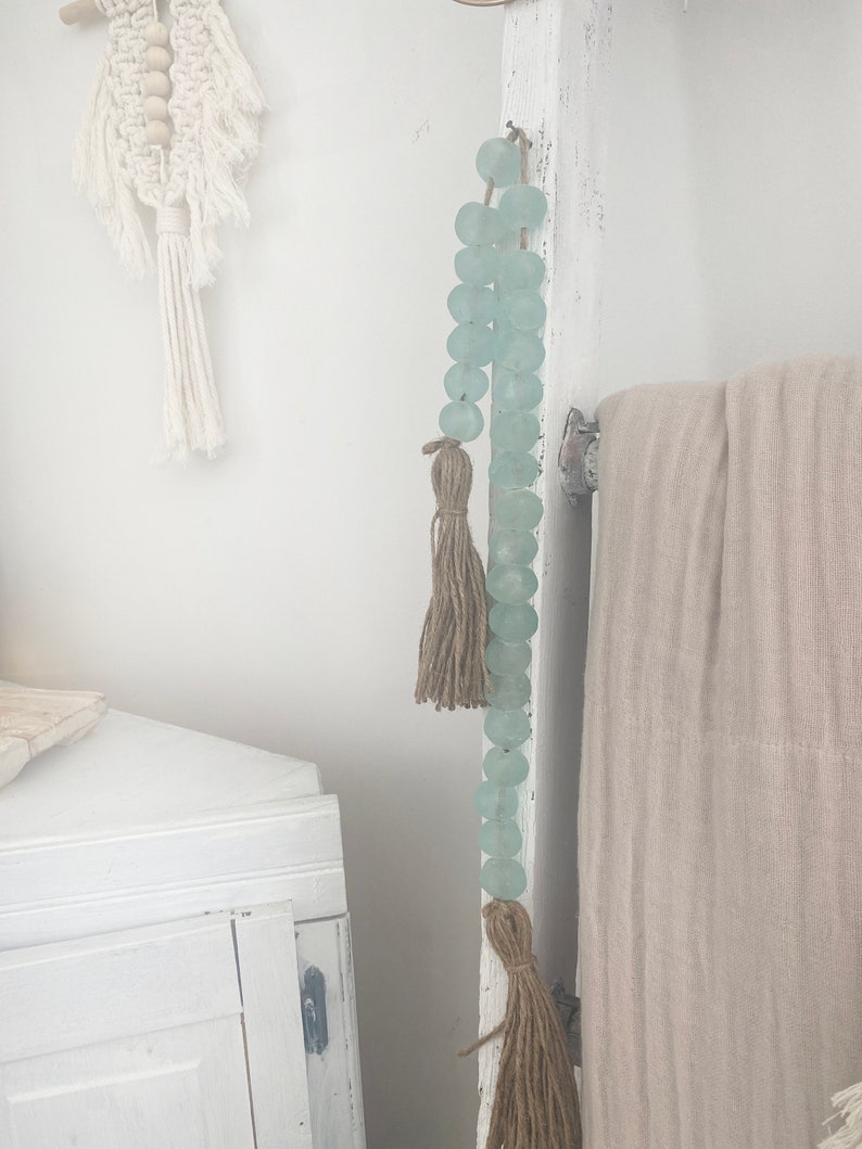Op de afbeelding: Een lichtblauwe glazen kralenkrans met bruine kwastjes die aan een witte houten ladder hangt. De krans is gemaakt van ronde kralen en heeft een rustieke, bohemian stijl.