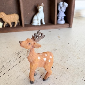 May include: A small, orange deer figurine with white spots and detailed antlers stands in front of a wooden display case. Other miniature animal figurines, including a sheep and a dog, are visible in the background.