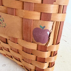 May include: Close-up of a woven basket with a rectangular shape. The basket is made of light brown and reddish-brown woven strips. A purple apple with a green stem is painted on the side of the basket. The basket appears to be handmade.