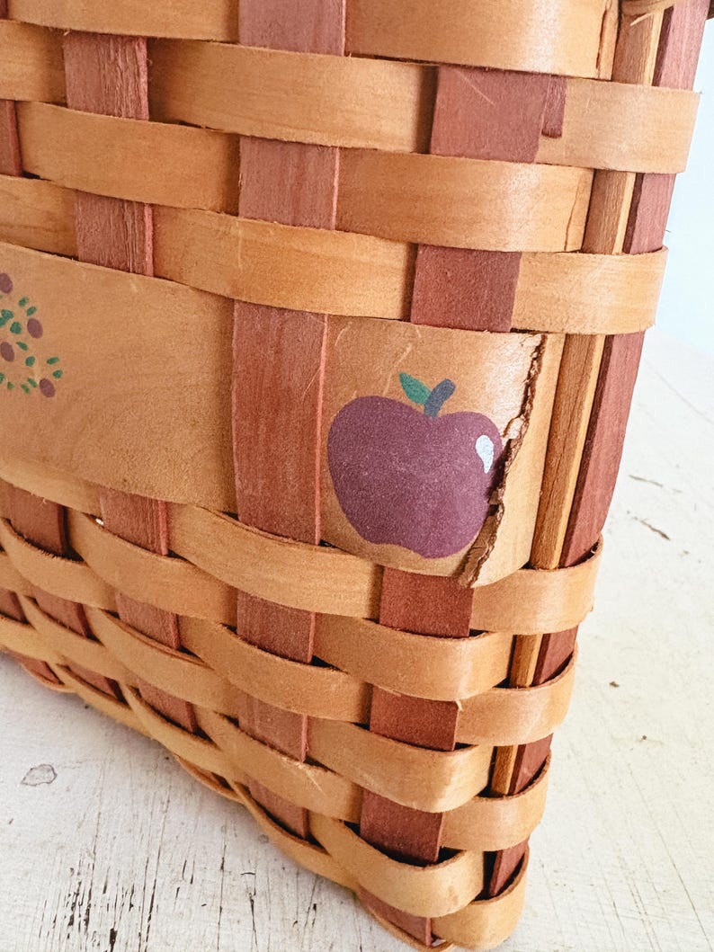 May include: Close-up of a woven basket with a rectangular shape. The basket is made of light brown and reddish-brown woven strips. A purple apple with a green stem is painted on the side of the basket. The basket appears to be handmade.