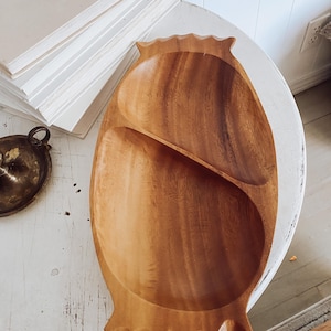 May include: A wooden, oval-shaped serving bowl with two compartments. The bowl is light brown with a natural wood grain and has a decorative, scalloped edge. The bowl is on a white, distressed table.