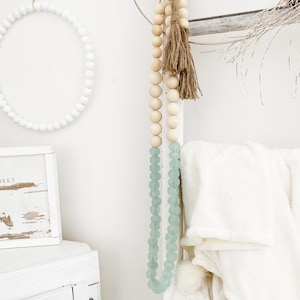 May include: A white wooden bead garland with a light blue glass bead garland hanging from it. The garlands are hanging from a white wall with a white wooden cabinet and a white wooden wreath in the background.