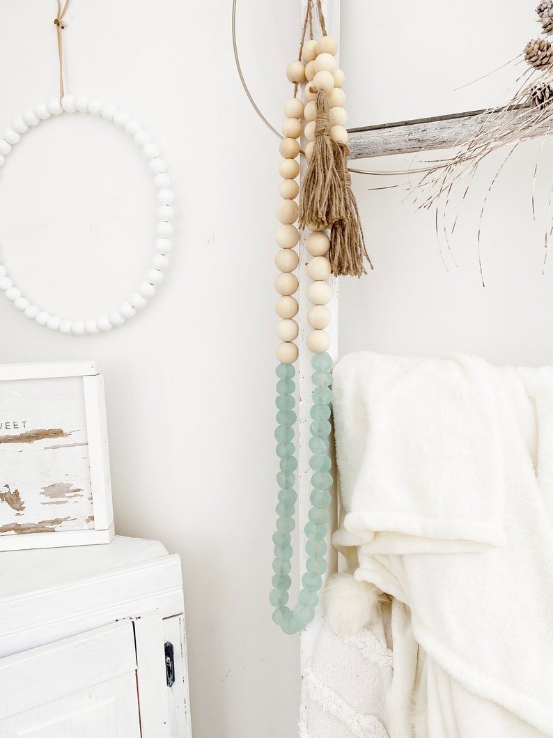May include: A white wooden bead garland with a light blue glass bead garland hanging from it. The garlands are hanging from a white wall with a white wooden cabinet and a white wooden wreath in the background.