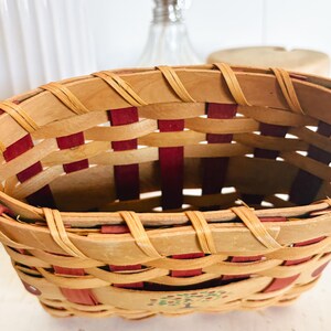 May include: Oval-shaped woven basket with a natural wood and red color scheme. The basket features a woven design with red accents and a painted tree design on the side. The basket is made of wood.