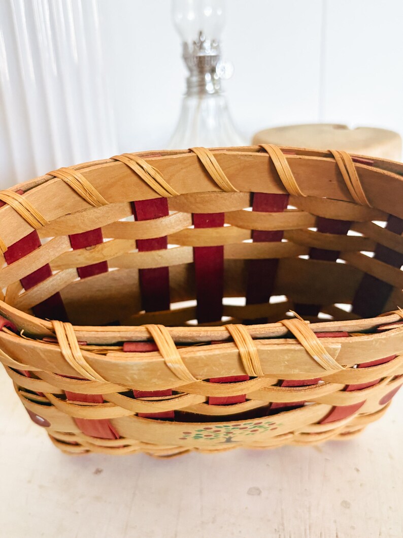 May include: Oval-shaped woven basket with a natural wood and red color scheme. The basket features a woven design with red accents and a painted tree design on the side. The basket is made of wood.