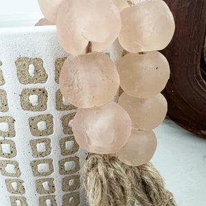 May include: A strand of pale pink glass beads with a natural fibre tassel. The beads are round and smooth, and the tassel is made of a natural fibre like jute or hemp.