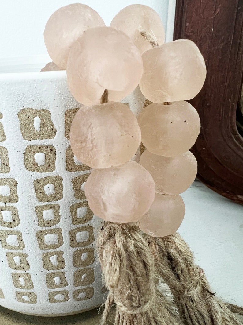 May include: A strand of pale pink glass beads with a natural fibre tassel. The beads are round and smooth, and the tassel is made of a natural fibre like jute or hemp.
