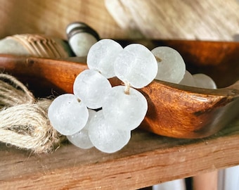 May include: A strand of translucent, frosted white glass beads strung together on a natural tan jute twine. The beads have a rounded, slightly irregular shape and a matte texture, creating a cluster that rests against a smooth, dark brown wooden bowl.