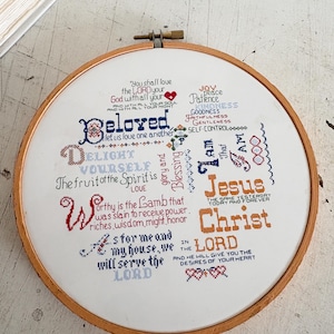May include: A cross-stitch embroidery hoop with religious text. The text includes phrases like "Beloved, let us love one another" and "Jesus Christ." The embroidery uses blue, red, and orange thread on white fabric, framed by a wooden hoop.