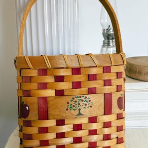 May include: Woven basket with a wooden handle. The basket features a natural wood color with red accents. The front of the basket has a painted apple tree design and two red apples. A charming decorative piece.