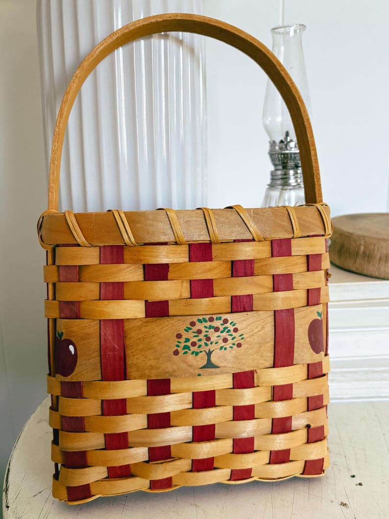 May include: Woven basket with a wooden handle. The basket features a natural wood color with red accents. The front of the basket has a painted apple tree design and two red apples. A charming decorative piece.