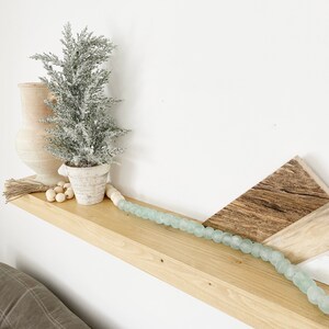 May include: A wooden shelf with a small, frosted artificial Christmas tree, a white ceramic vase, and a string of light blue glass beads. The shelf is mounted on a white wall.