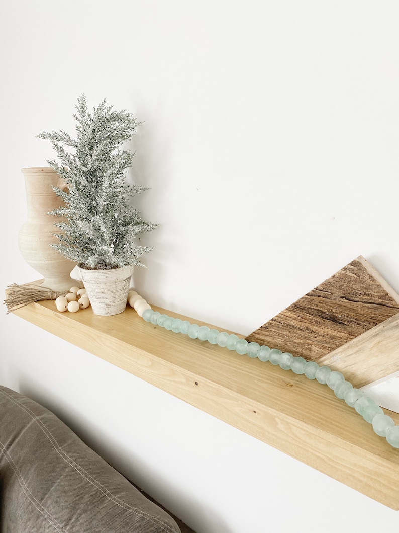 May include: A wooden shelf with a small, frosted artificial Christmas tree, a white ceramic vase, and a string of light blue glass beads. The shelf is mounted on a white wall.
