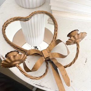 May include: A gold-toned metal candle holder featuring a heart-shaped mirror frame, a central decorative bow, and two twisted arms that extend outward to support scalloped candle cups. The finish has a distressed, weathered appearance.