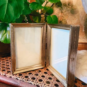 May include: A double-hinged, rectangular picture frame with a metallic gold finish. The frame features two vertical openings for photos, connected by a central hinge, and includes decorative etched detailing along the outer side edges.