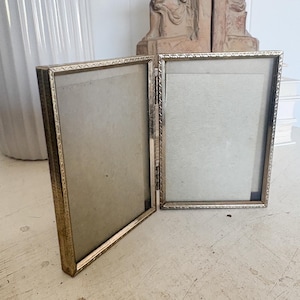May include: A vintage, hinged, double-sided photo frame. The frame is made of a gold-colored metal with a decorative border. The frame is open, revealing the space for two photos. The frame is resting on a distressed, white surface.