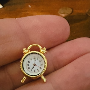 May include: A miniature gold-colored alarm clock with a white face and black numerals. The clock has a small handle at the top and two bells. The hands point to approximately 10:07. The clock is a decorative item.
