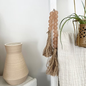 May include: A white wooden ladder with a blush pink beaded garland with tassels hanging from it. A white throw blanket is draped over the ladder. A potted plant is on the top shelf of the ladder.