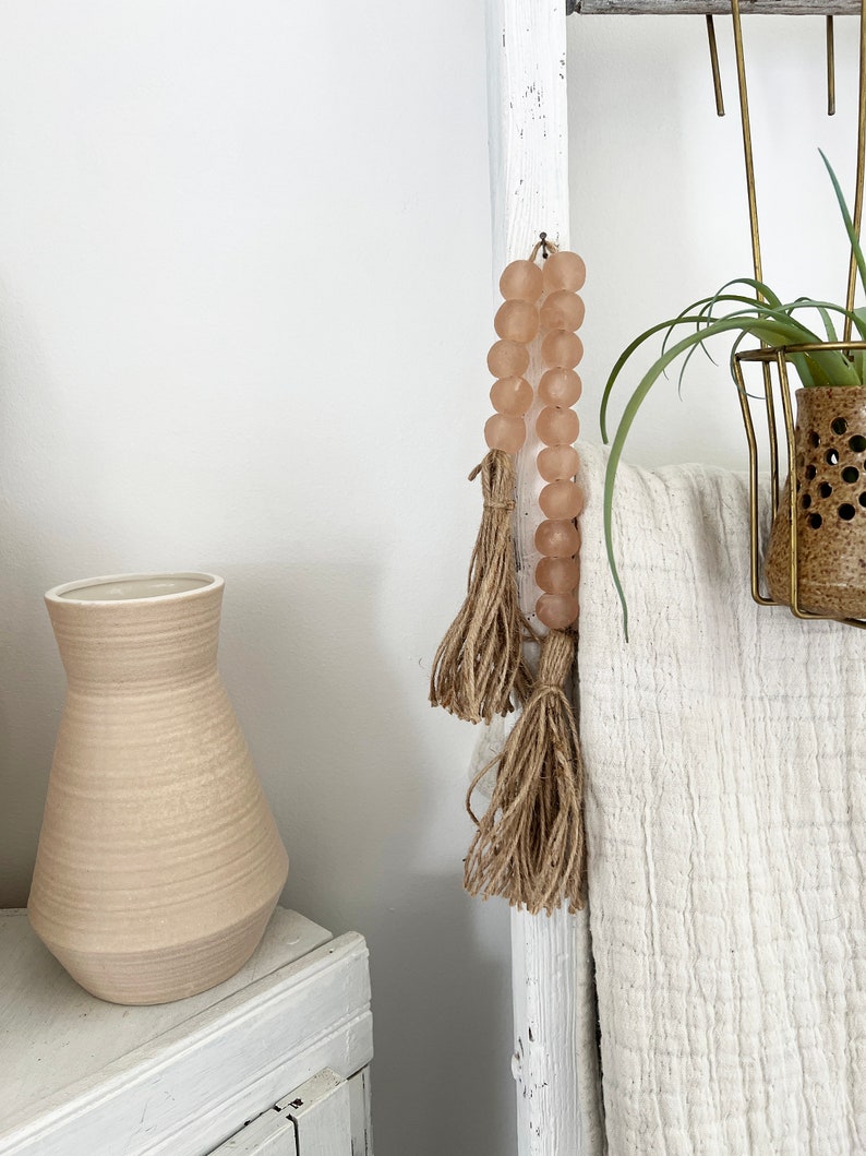 May include: A white wooden ladder with a blush pink beaded garland with tassels hanging from it. A white throw blanket is draped over the ladder. A potted plant is on the top shelf of the ladder.