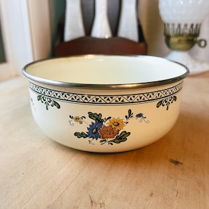May include: Cream-colored enamel bowl with a decorative floral pattern in blue, orange, and yellow. The bowl has a silver rim and a black and green geometric border. A vintage kitchenware item.