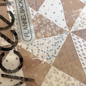 May include: A flannel back table cover featuring a geometric pattern of triangles in tan, white, and blue. The design includes small floral and leaf motifs. The packaging text reads FLANNEL BACK TABLE COVER in black capital letters.