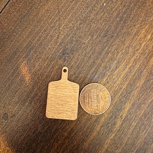 May include: A small, wooden cutting board with a handle, next to a penny. The cutting board is rectangular with a rounded handle and a hole for hanging. The penny shows the profile of Abraham Lincoln.