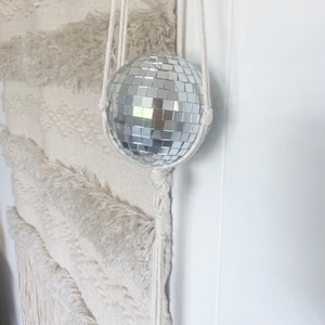 May include: A silver disco ball hanging from a white macrame plant hanger.