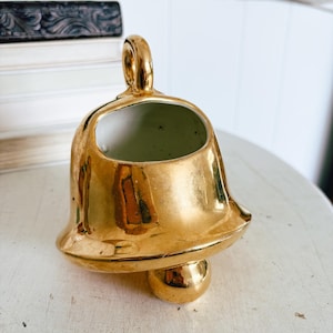 May include: A gold-colored bell-shaped ceramic planter with a handle. The planter has a wide opening and sits on a small, round base. The surface has a shiny, metallic finish.