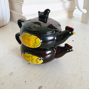 May include: A stacked, ceramic hen-shaped teapot and creamer set. The teapot is black with a small black bird on the lid. The creamer has a red comb and yellow wing accents. The set is on a white surface.