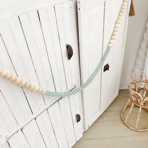 May include: White wooden cabinet with two doors and beaded garland draped across the front. The garland is made of light brown and light blue beads.